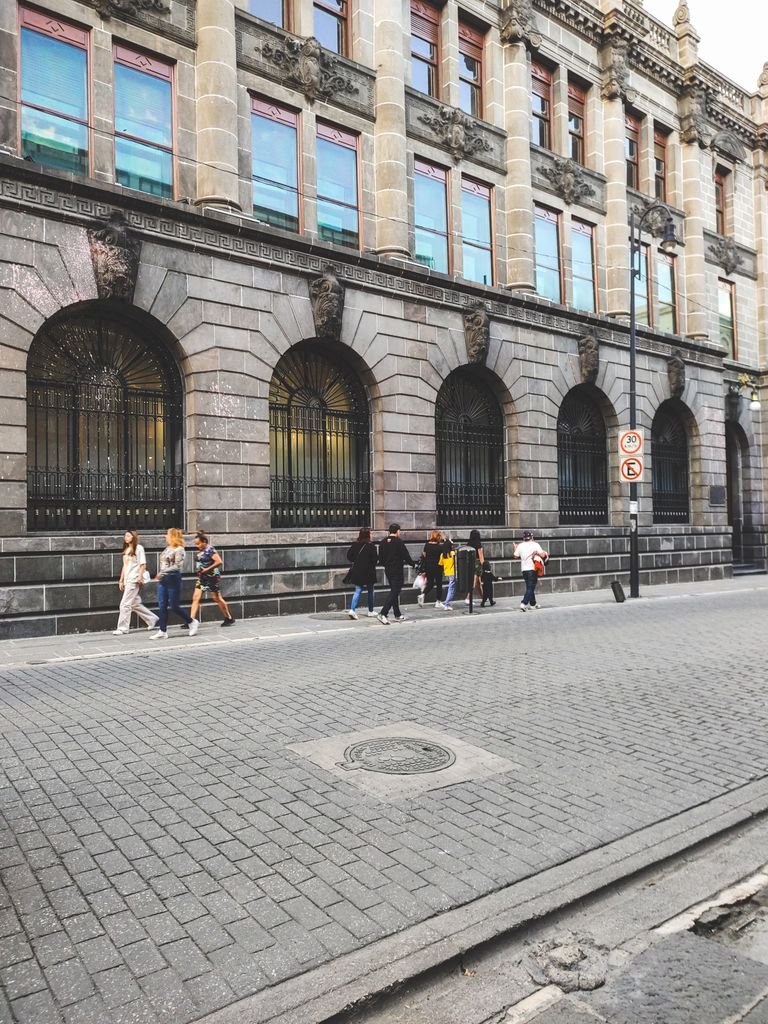 People walking by a historical stone building with arched windows on a quiet street.